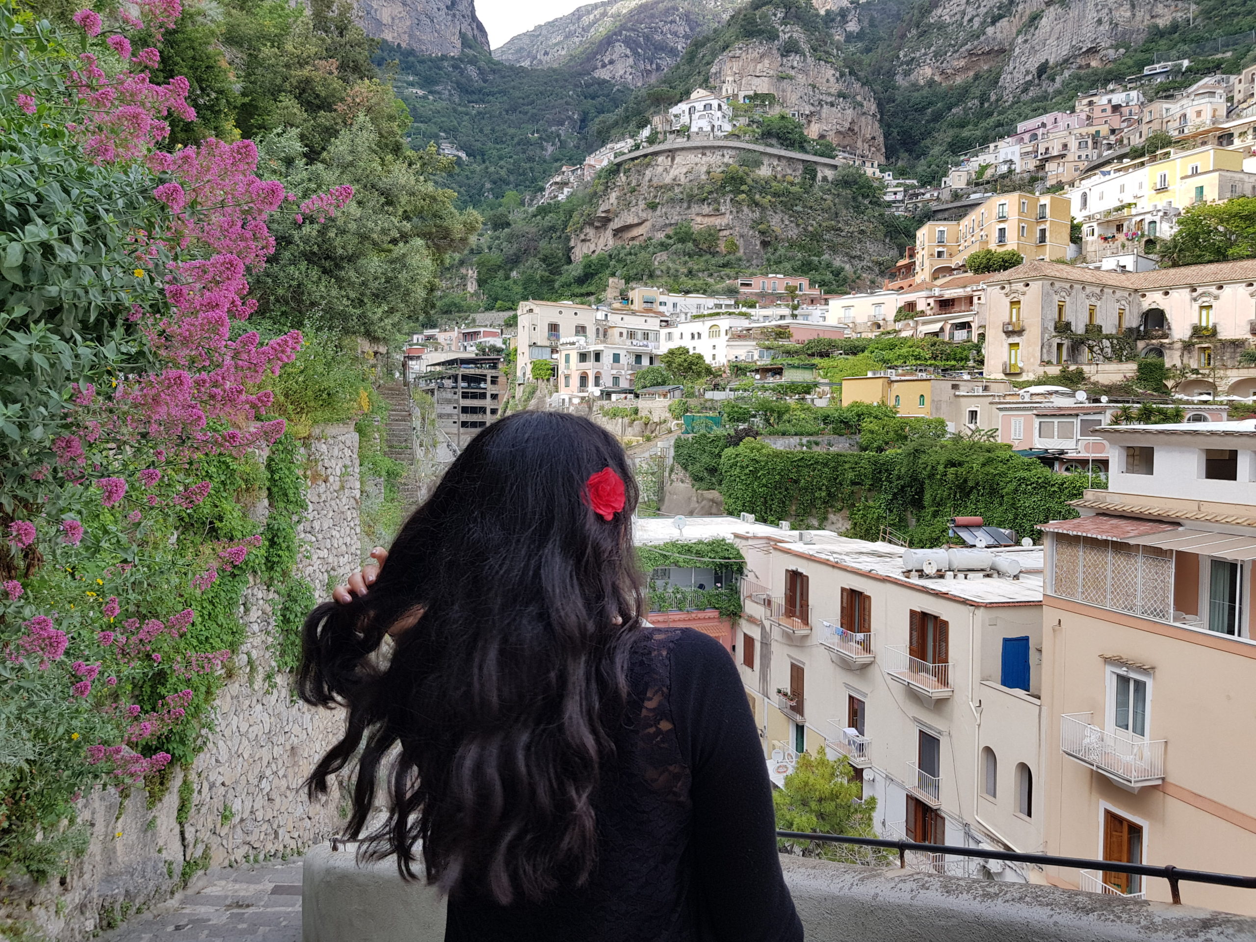 Woman is standing with her back to the camera with her dark hair loose and a red flower on the right side of her hair. She is flicking her hair over her left shoulder. In the background is a beautiful view of the hills and architecture of Positano, Amalfi Coast, Italy.
