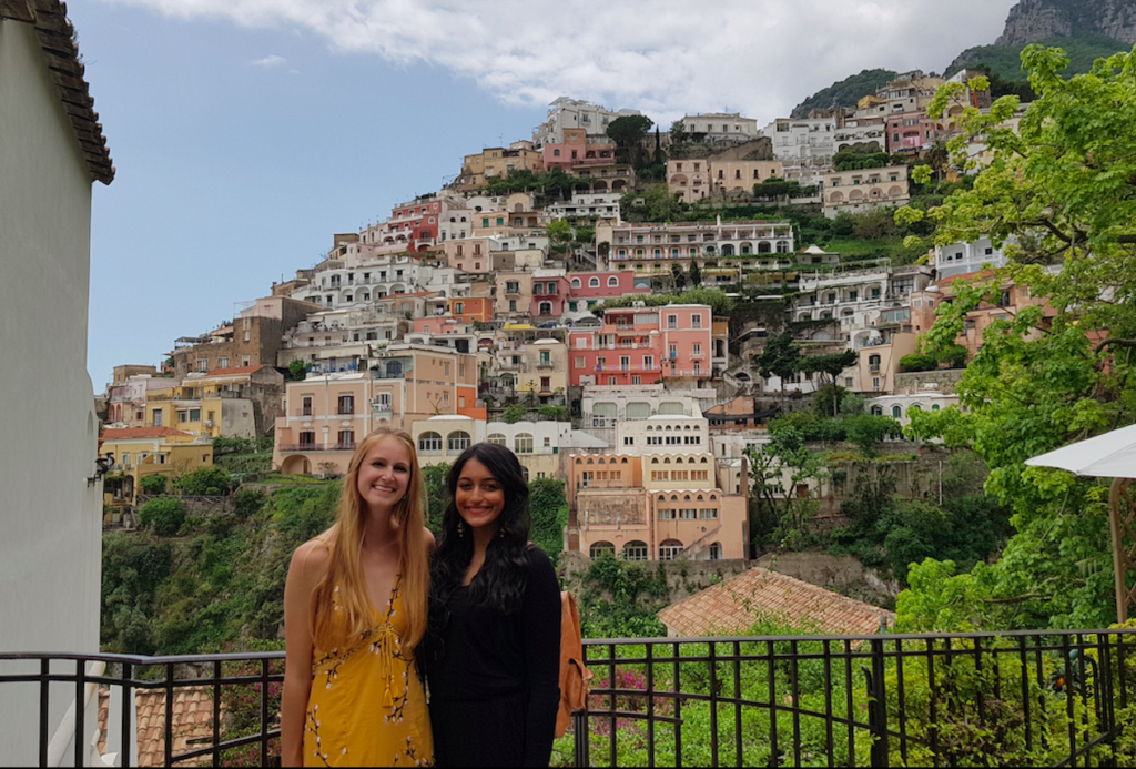 Two women are smiling into the camera, one is wearing a yellow sundress and the other iis wearing a back jumpsuit and cardigan. Behind them are Positano's picturesque hills and buildings.