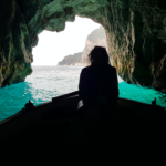 Silhouette of a woman sat on a boat inside a cave. In the background you can see the opening of the cave and turquoise blue water.